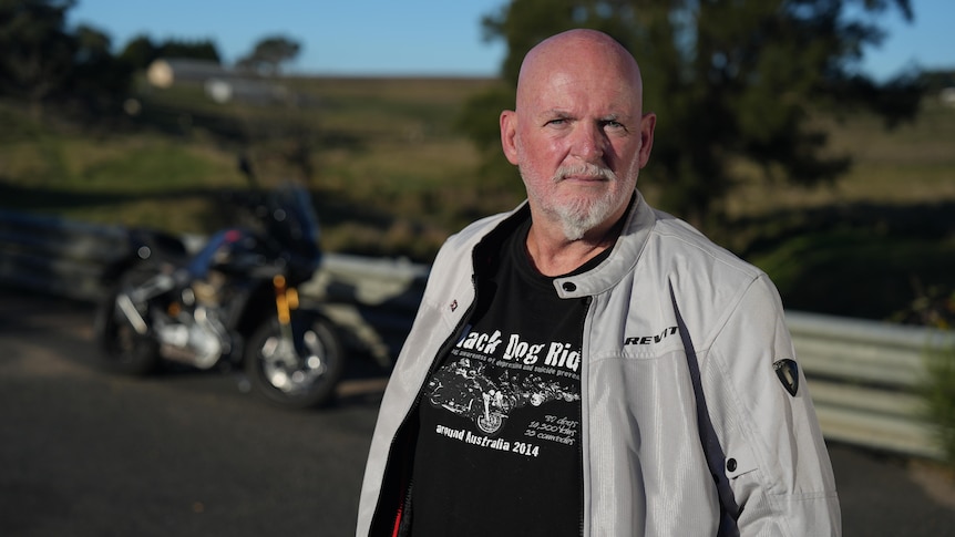 A middle aged bald white man standing on a country road in front of a parked motorbike.