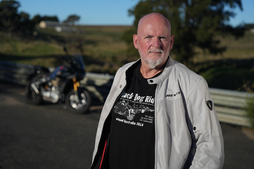 A middle aged bald white man standing on a country road in front of a parked motorbike.