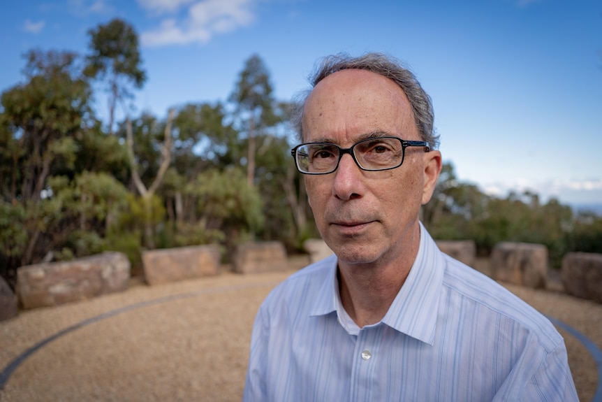 A man wearing a light blue shirt, glasses looks at the camera, green trees behind.