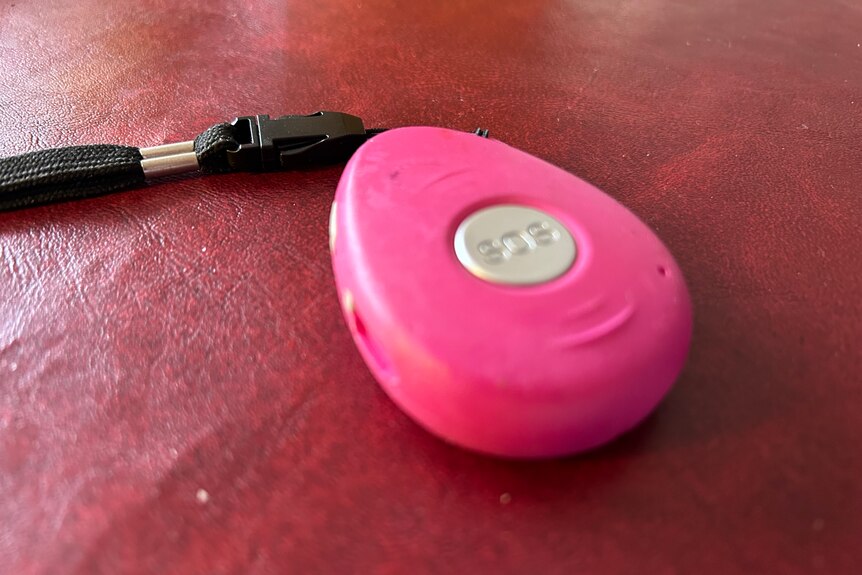 A pink medical alert pendant with a silver SOS button and a black strap sitting on a red table top.