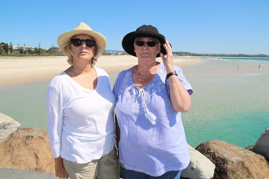 Two women stand side by side at a rocky outcrop overlooking the beach