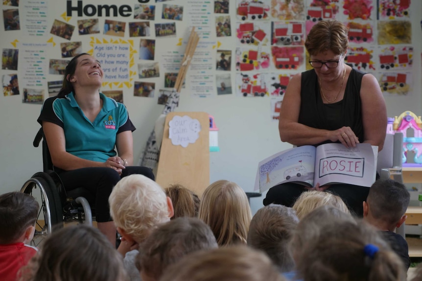 A lady sits in a wheelchair laughing, while another lady reads a picture book to a group of young children.