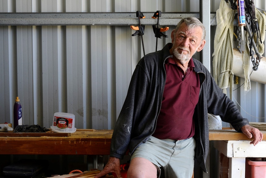 A man stands on one leg leaning on a wooden cray trap