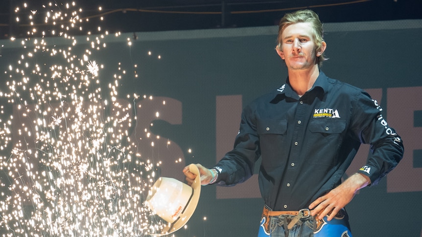 A young cowboy stands next to a shower of sparks spitting out of some kind of Roman candle.