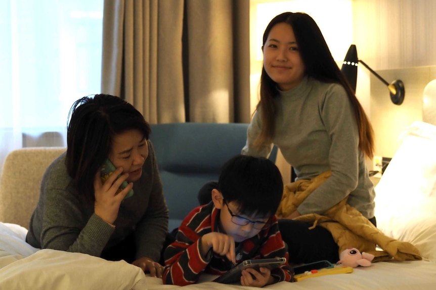 A family lie on a hotel bed, with one woman talking on a mobile phone while a child plays on a tablet