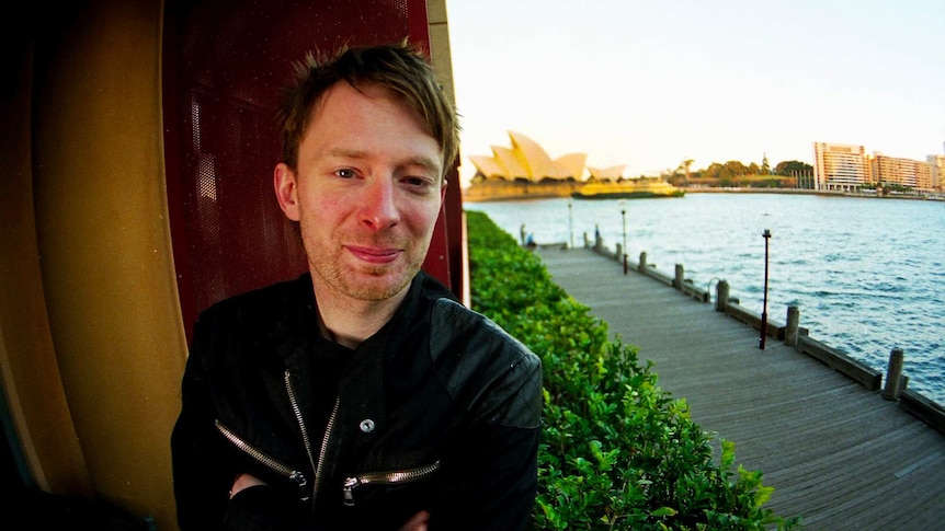 Radiohead's Thom Yorke smiling with his arms crossed. Sydney harbour and Sydney Opera House are in the background