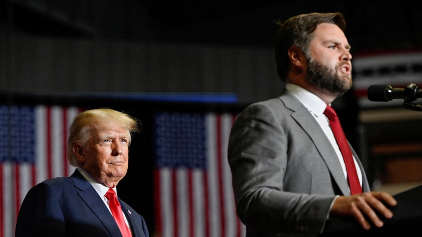 JD Vance speaks at a lectern as Donald Trump stands behind watching, with two US flags