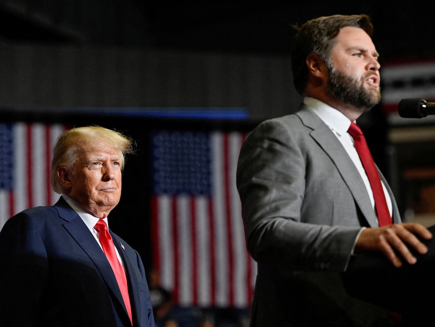 JD Vance speaks at a lectern as Donald Trump stands behind watching, with two US flags