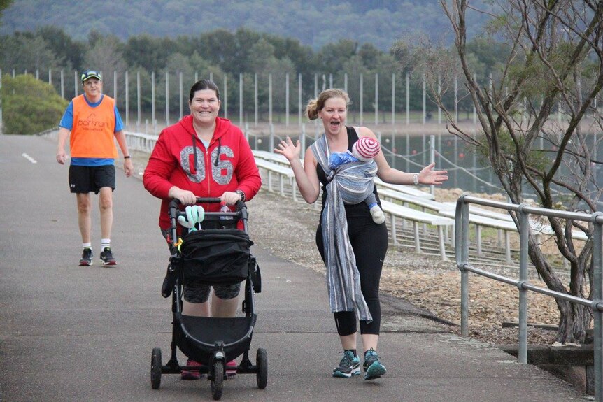Two women walk on a concrete path, one pushes a pram and the other has a baby in a carrier.