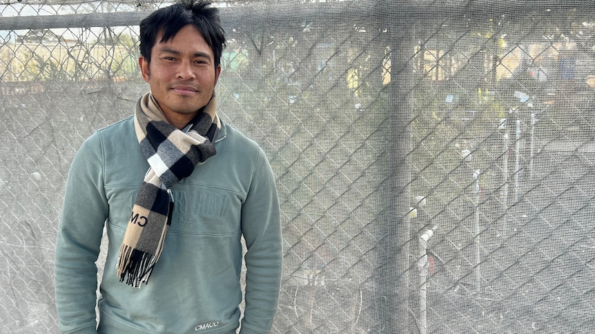 A man in a green jumper standing in front of a chain link fence.