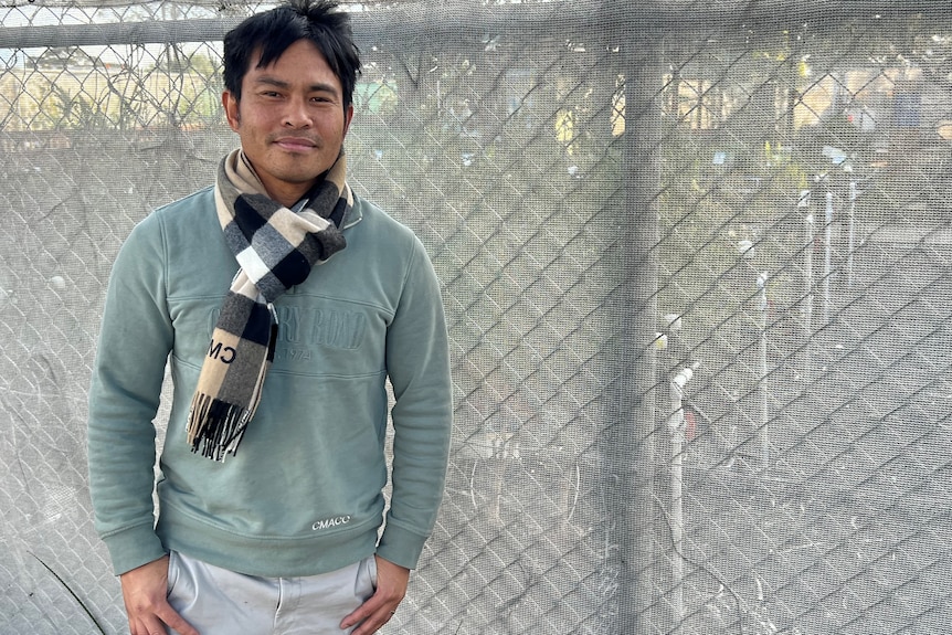 Wide shot of a man in a green jumper standing to left of frame in front of a chain link fence 