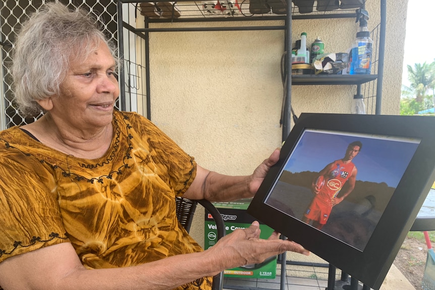 A woman in an orange shirt with gray hair smiles at a photo of a young man wearing an AFL uniform.