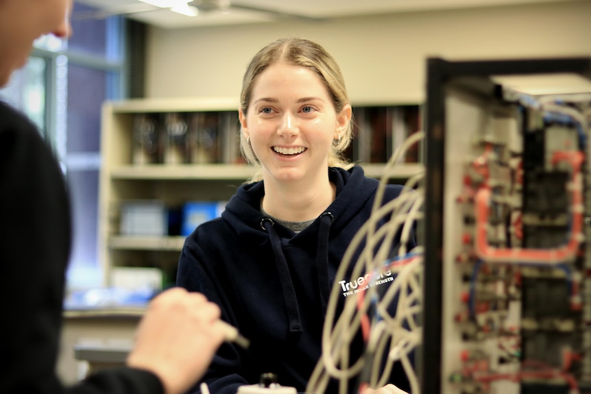 Lauren Blasi smiles, wearing a dark blue hoodie and behind a panel with cords coming out of it.