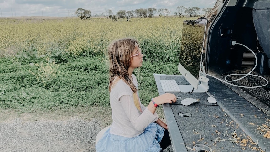 A young gild sits on a still at the back of a ute looking at a computer