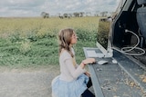 A young gild sits on a still at the back of a ute looking at a computer