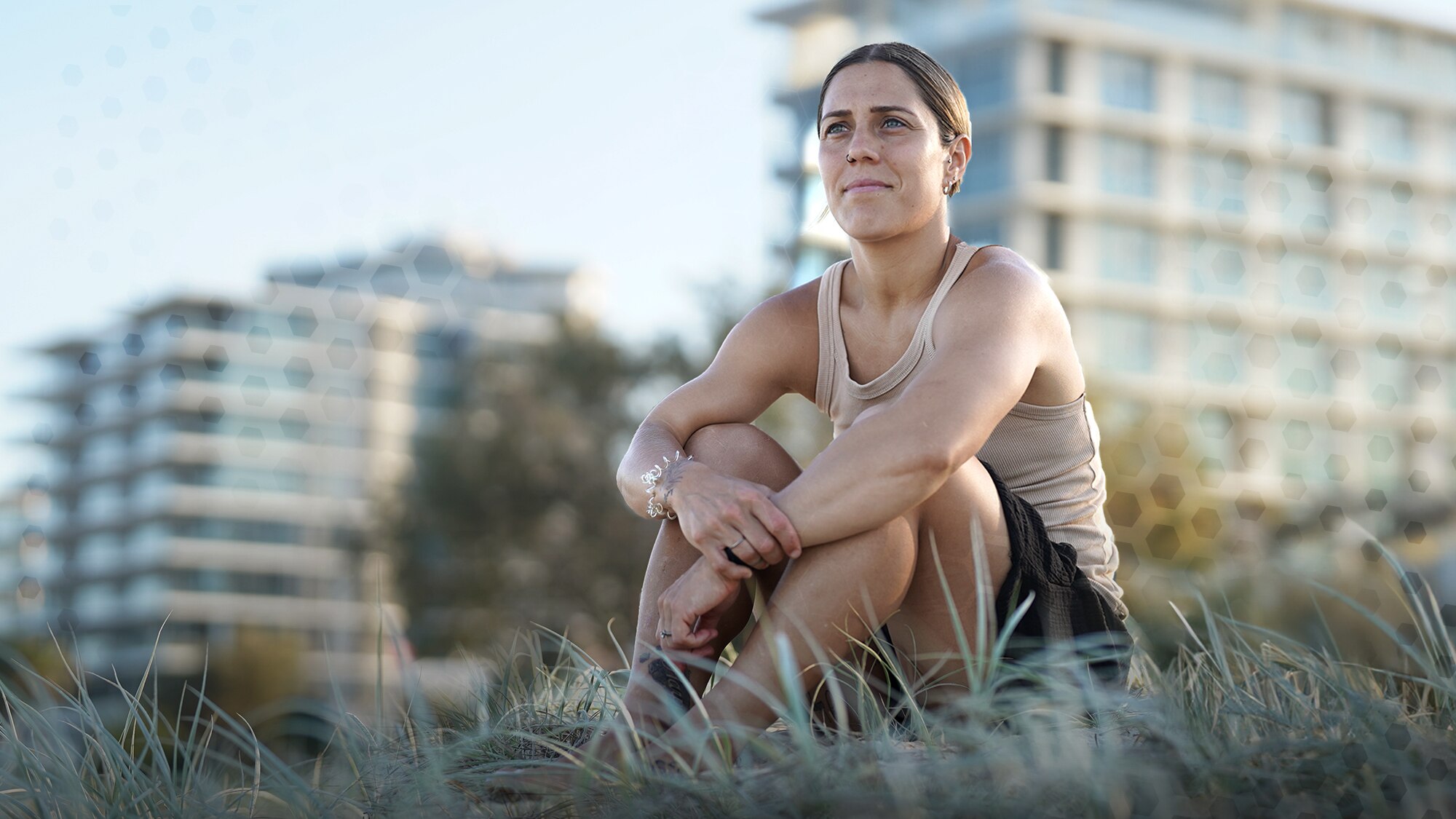 Woman in a beige singlet sits on the dunes at the beach looking out to distance 