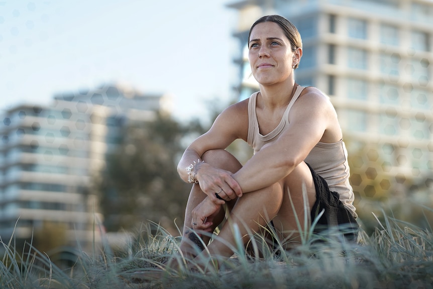 Woman in a beige singlet sits on the dunes at the beach looking out to distance 