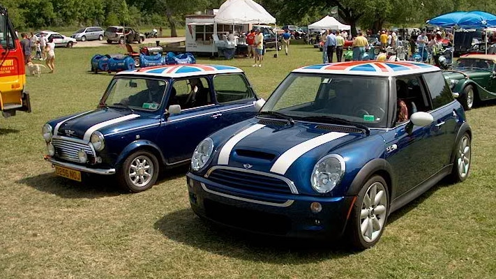An old and new Mini cooper sit next to each other on a green field which are both painted navy with a union jack on their roofs.