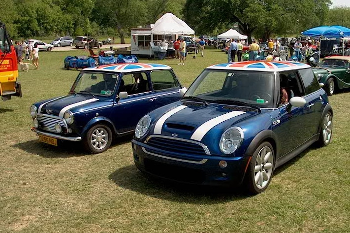 An old and new Mini cooper sit next to each other on a green field which are both painted navy with a union jack on their roofs.