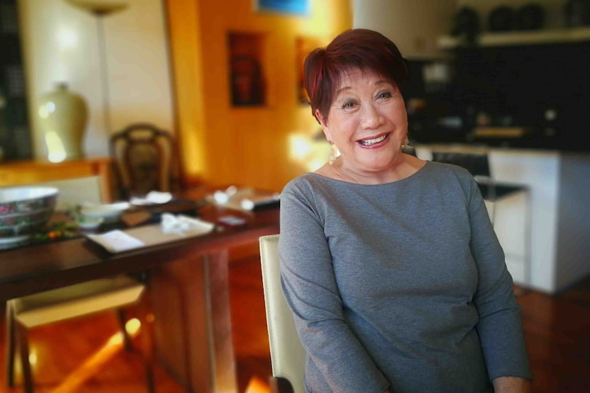 A smiling woman sits on a chair in front of a blurry, sunny indoor background.