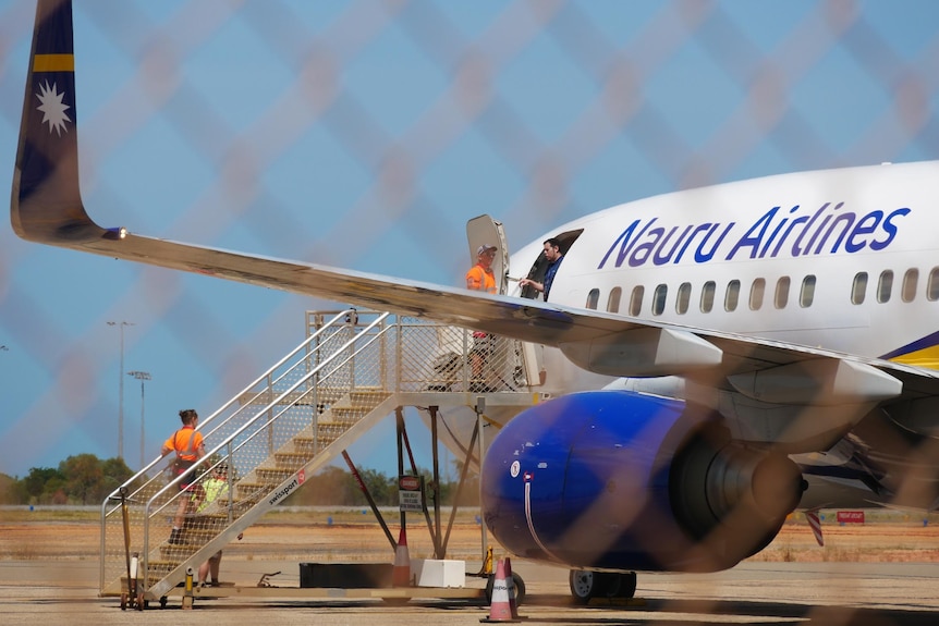 Workers in orange hi-vis boarding a plane photographed through a wired fence.