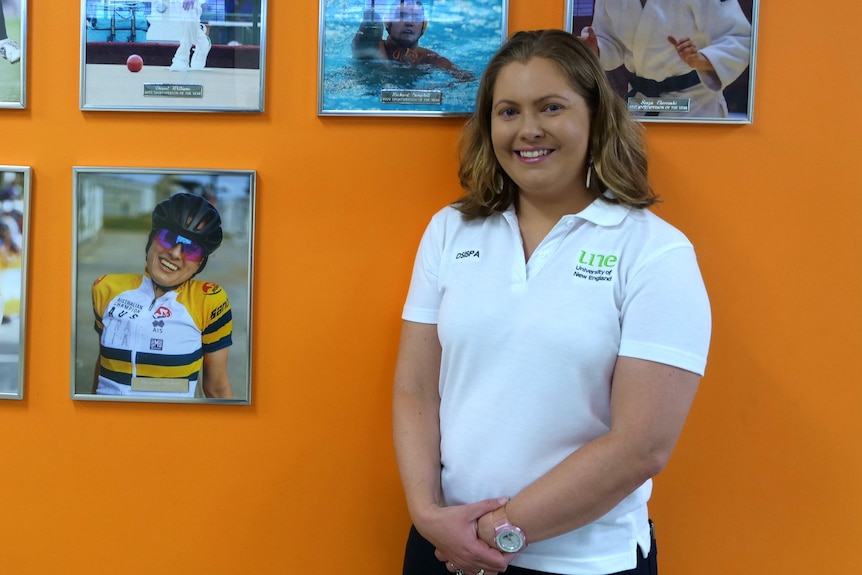 Dr. Kristy O'Neill stands in front of an orange wall with notable sporting identities from UNE behind her