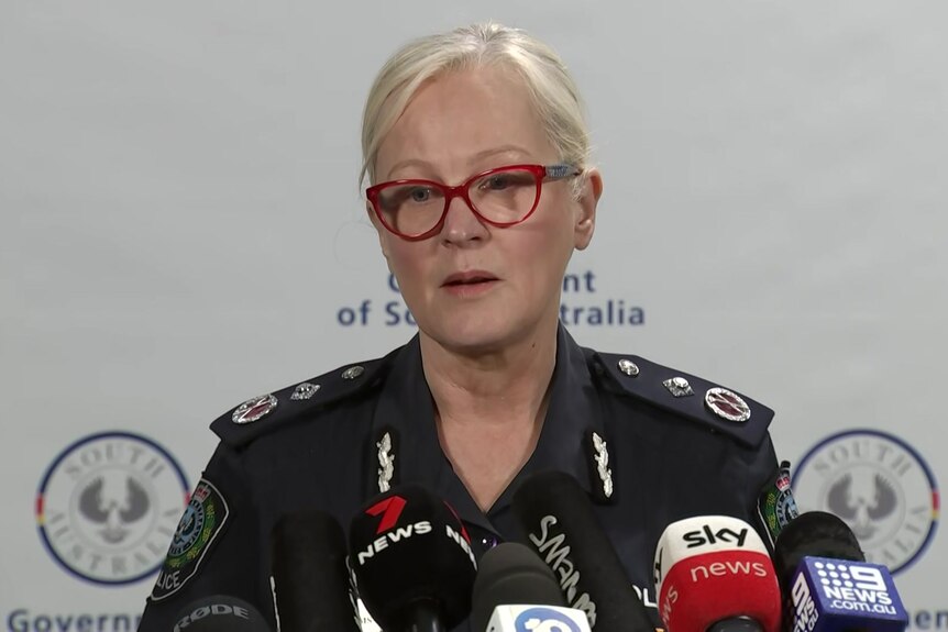 A female police officer with blonde hair speaks to microphones while standing in front of a white background