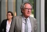 Geoffrey Rush, wearing a grey suit and tie, walks out of court.