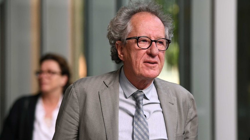 Geoffrey Rush, wearing a grey suit and tie, walks out of court.