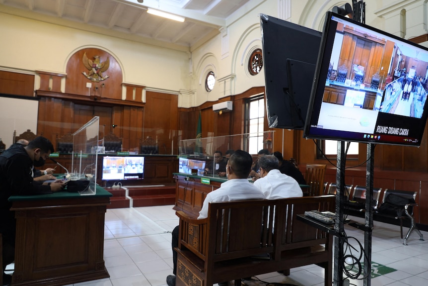 Two men in white business shirts sit in a wood-panelled courtroom, with clear perspex sheets and video screens around the room.