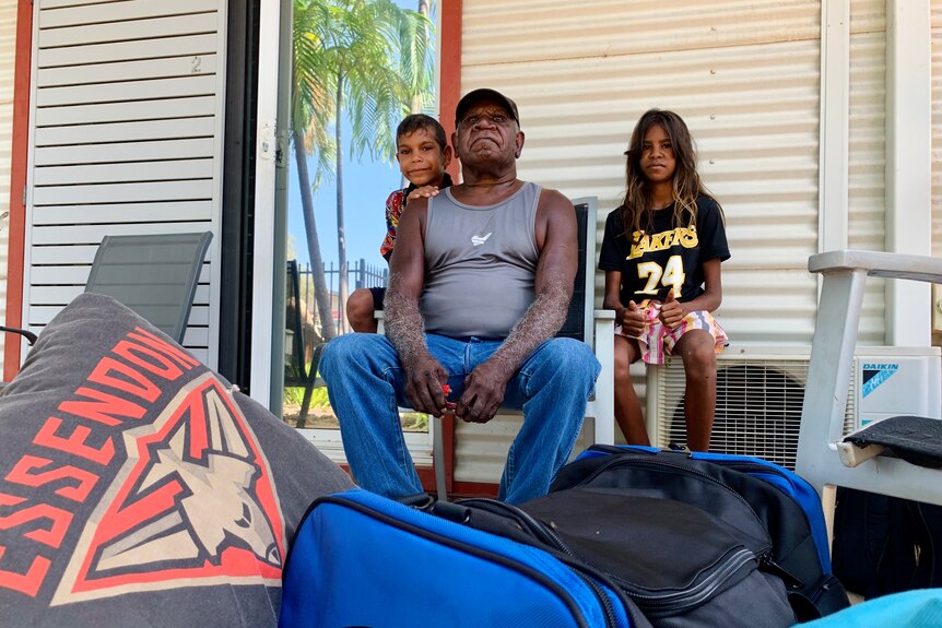 A man sits on a chair on a verandah flanked by two children with bags in the foreground.