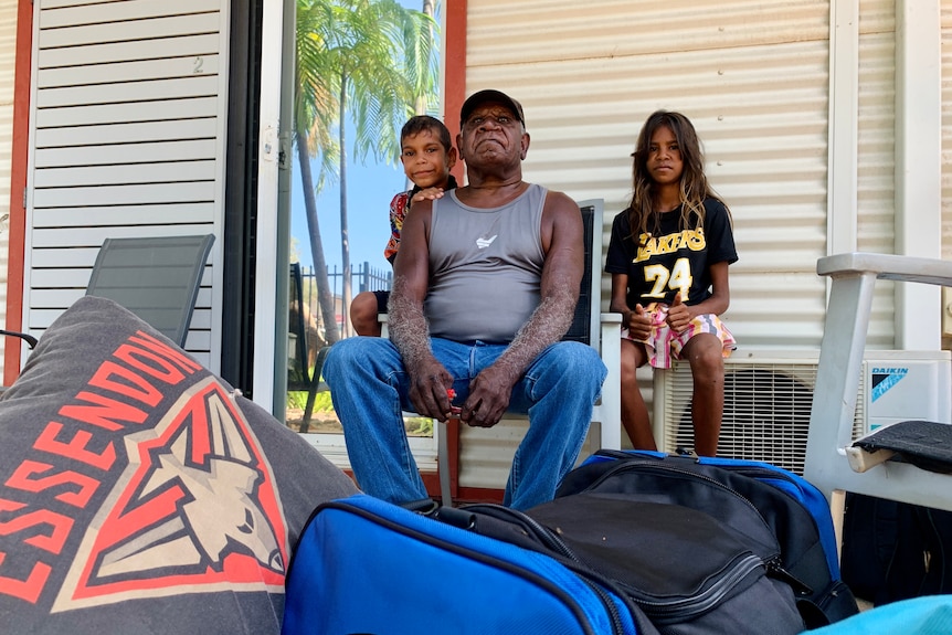 A man sits on a chair on a verandah flanked by two children with bags in the foreground.