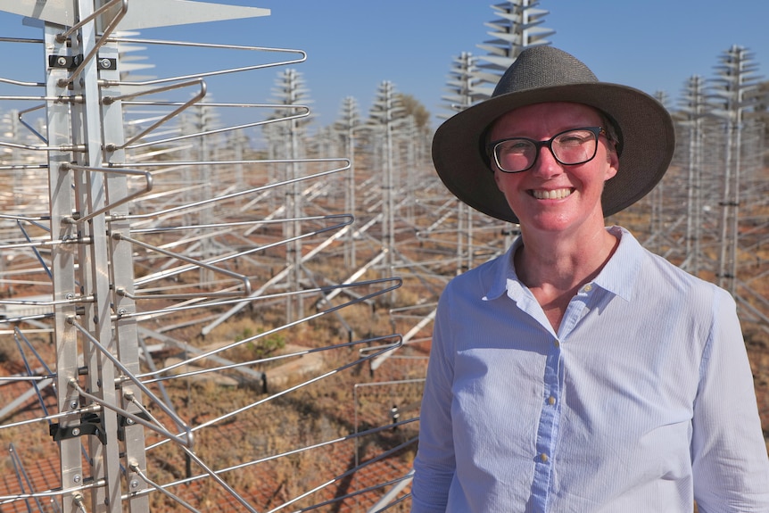 A woman in hat, sunglasses and buttoned shirt smiles at the camera with wire tree-like structures behind 
