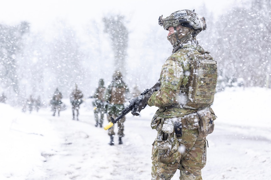 A soldier holding a firearm stands in snow with other soldiers walking in the background