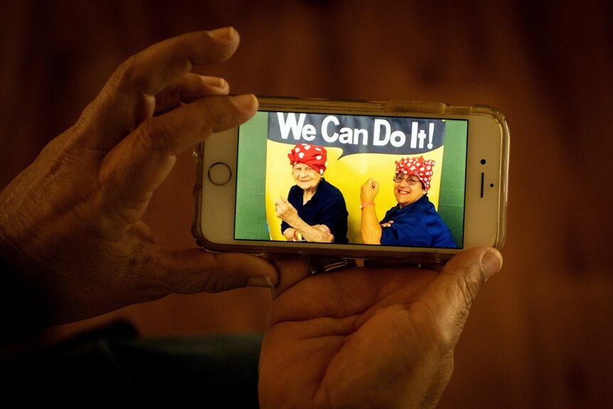 Two hands hold up an iphone. On the screen, an image of two women dressed as Rosie the Riveter.