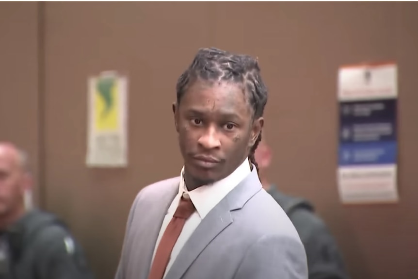 Young Thug, dressed in a grey suit and brown tie, is in a courtroom.
