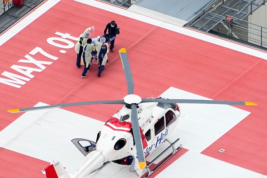 A medical helicopter sits on a red rooftop, people carry a patient into a building. 
