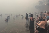 People stand in the water and on the edge of the beach as smoke fills the air.