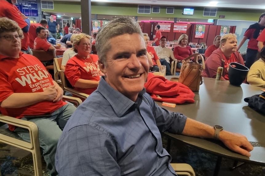 Smiling man in collared dress shirt, sits at a table, surrounded by people wearing red shirts