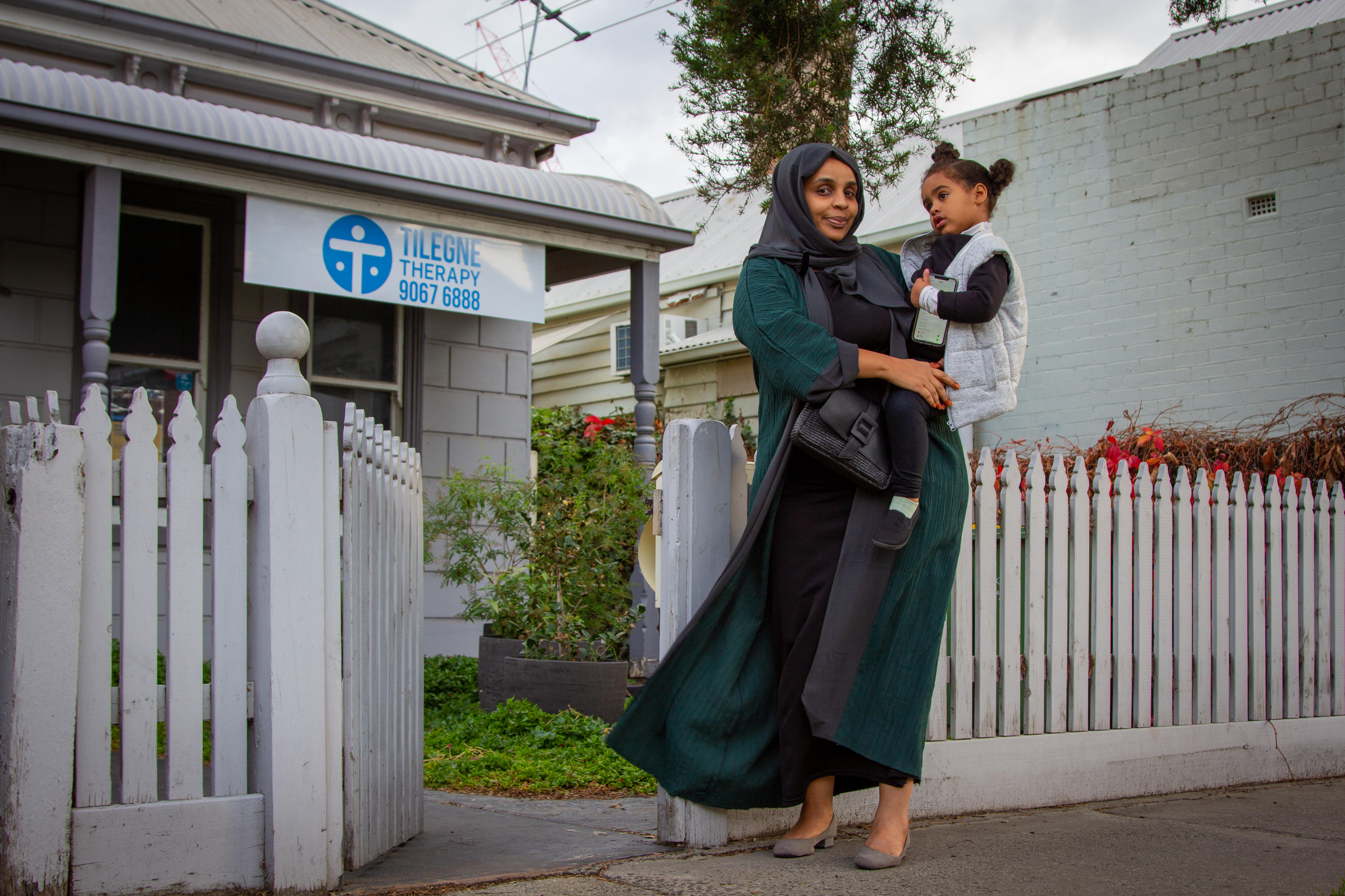 A woman holds her toddler in front of a house with a sign saying 'Tilegne Therapy'