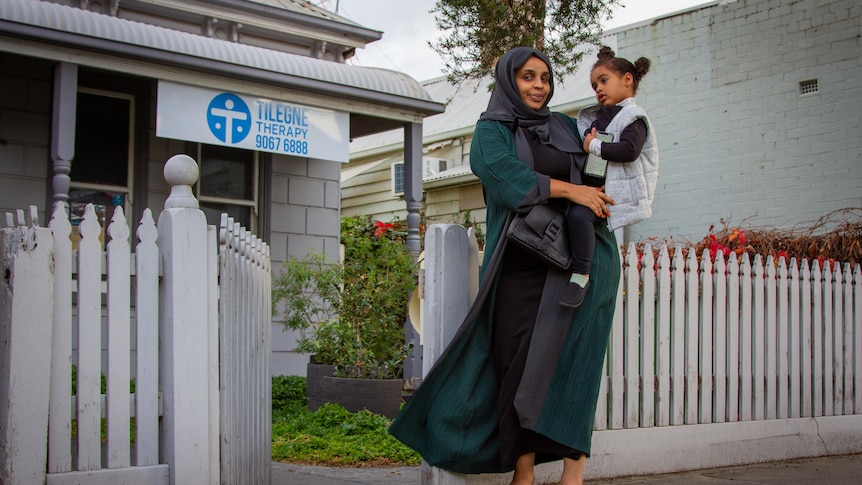 A woman holds her toddler in front of a house with a sign saying 'Tilegne Therapy'