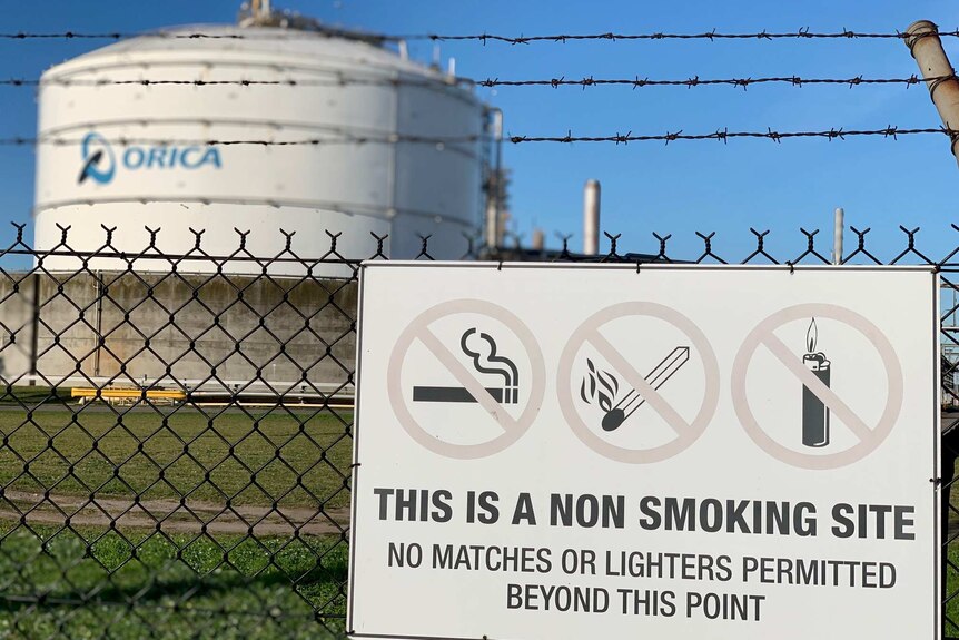 A sign on a perimeter wire fence warns of no smoking or flame on site, in the background is a very large tank with Orica logo