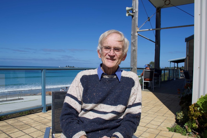An older gentleman with glasses sits near the ocean.