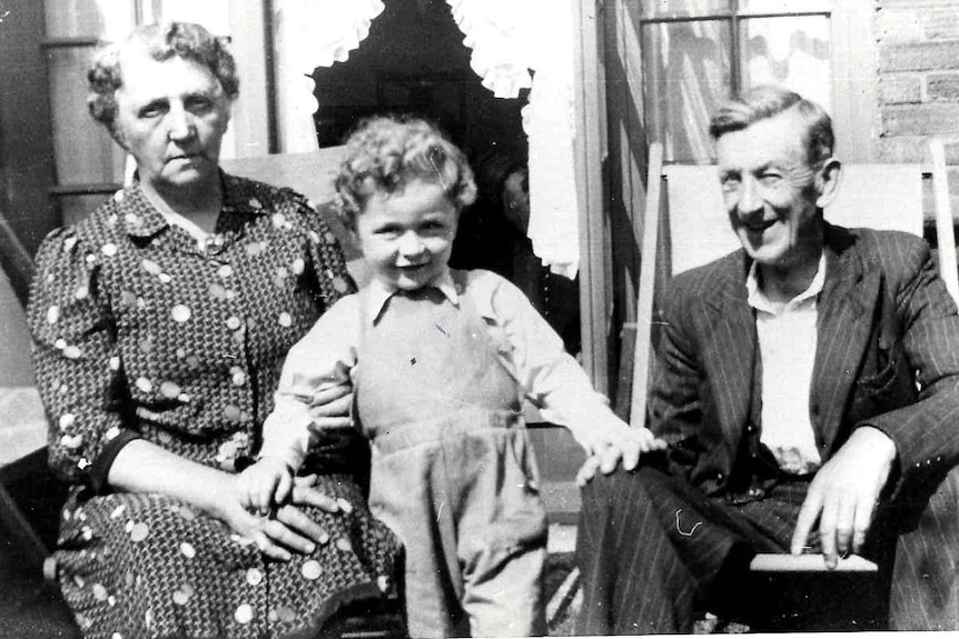 Black and white photo of a little boy standing between an elderly woman and man sitting on chairs.