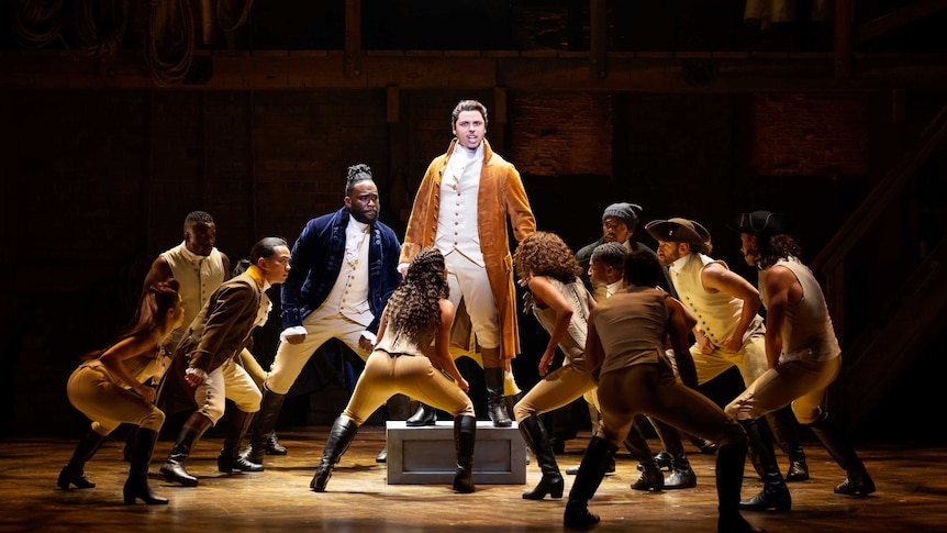 A scene from the musical Hamilton with the protagonist Alexander Hamilton standing on a box on a stage surrounded by the cast