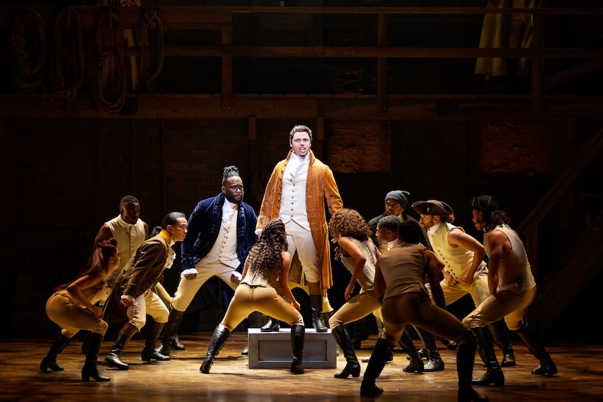 A scene from the musical Hamilton with the protagonist Alexander Hamilton standing on a box on a stage surrounded by the cast