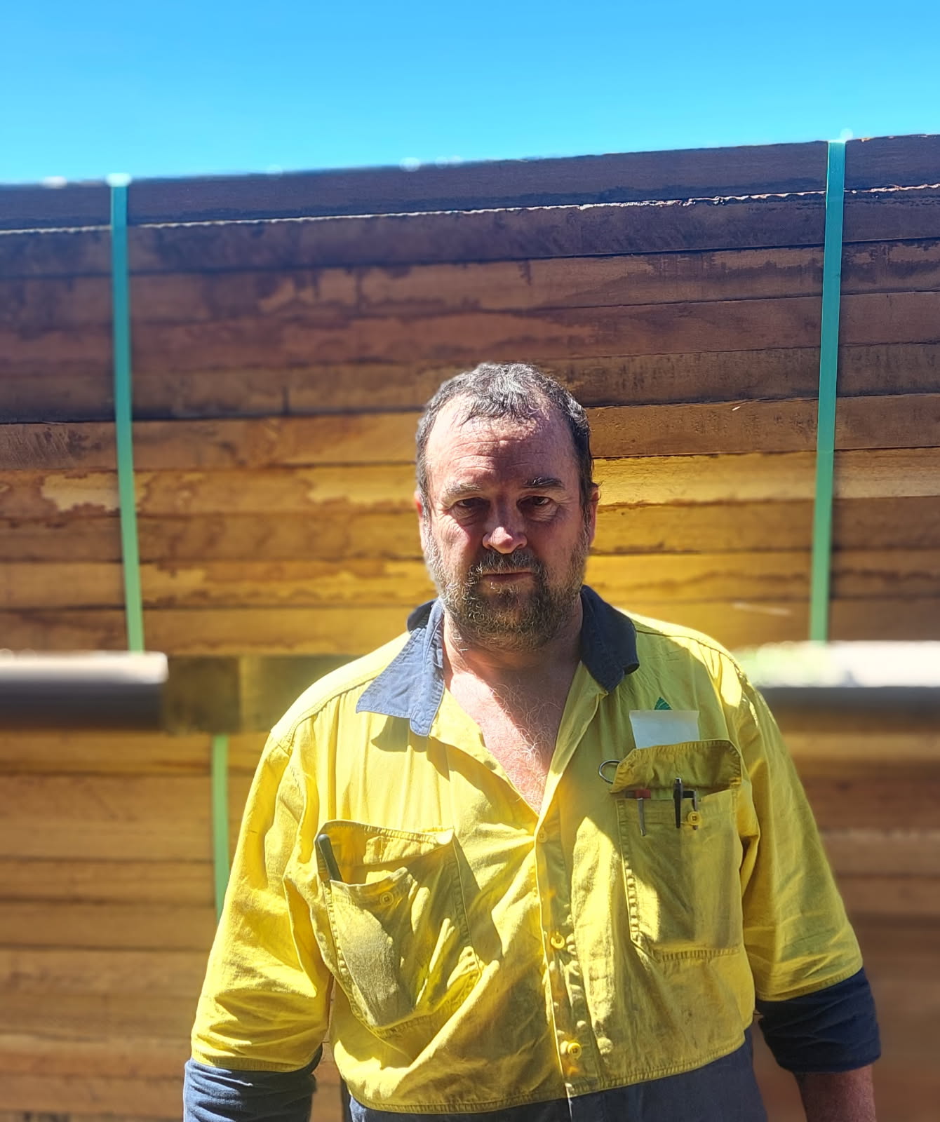 Homem de camisa de trabalho amarela em frente a madeira empilhada