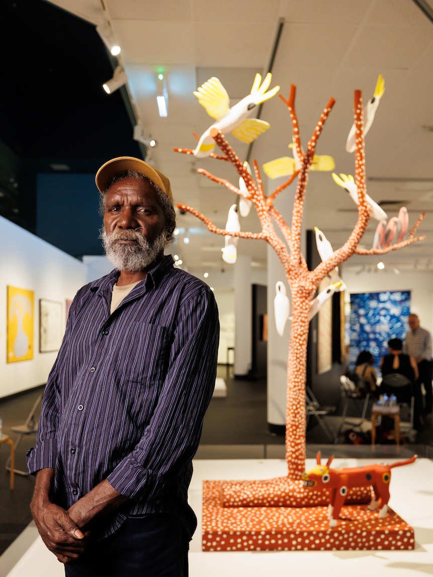 An Aboriginal man wearing a cap stands in front of a sculpture of a tree covered in birds