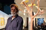 An Aboriginal man wearing a cap stands in front of a sculpture of a tree covered in birds
