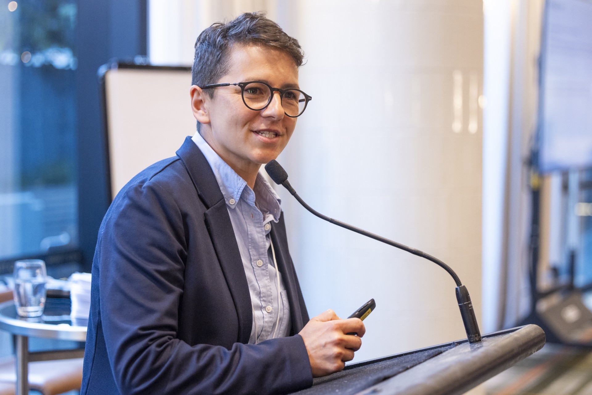 Lady with short hair and glasses, wearing a suit, standing at a lectern speaking and looking out toward her audience